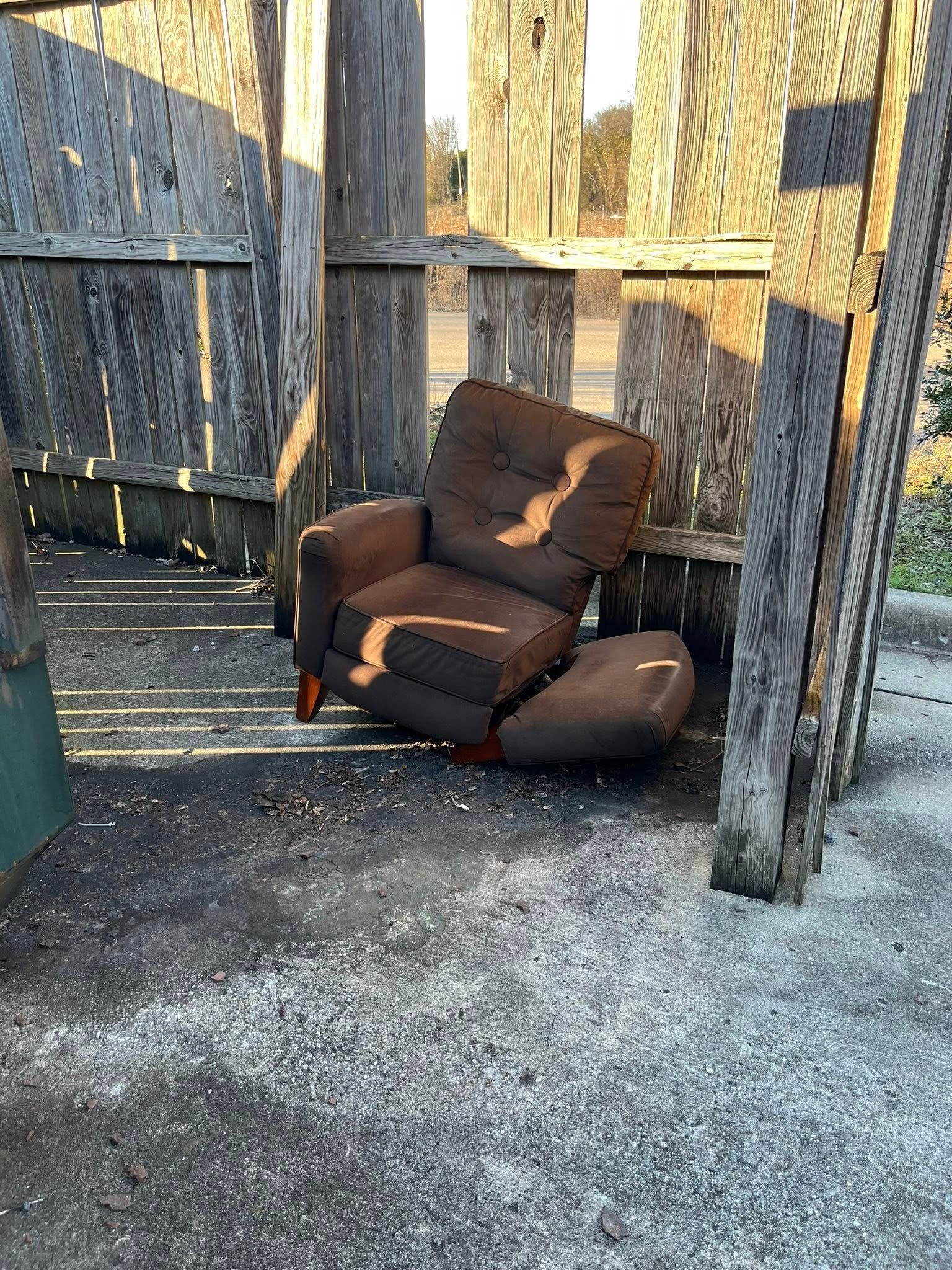 A worn, brown recliner sits abandoned in a dilapidated wooden structure with a dirt floor.