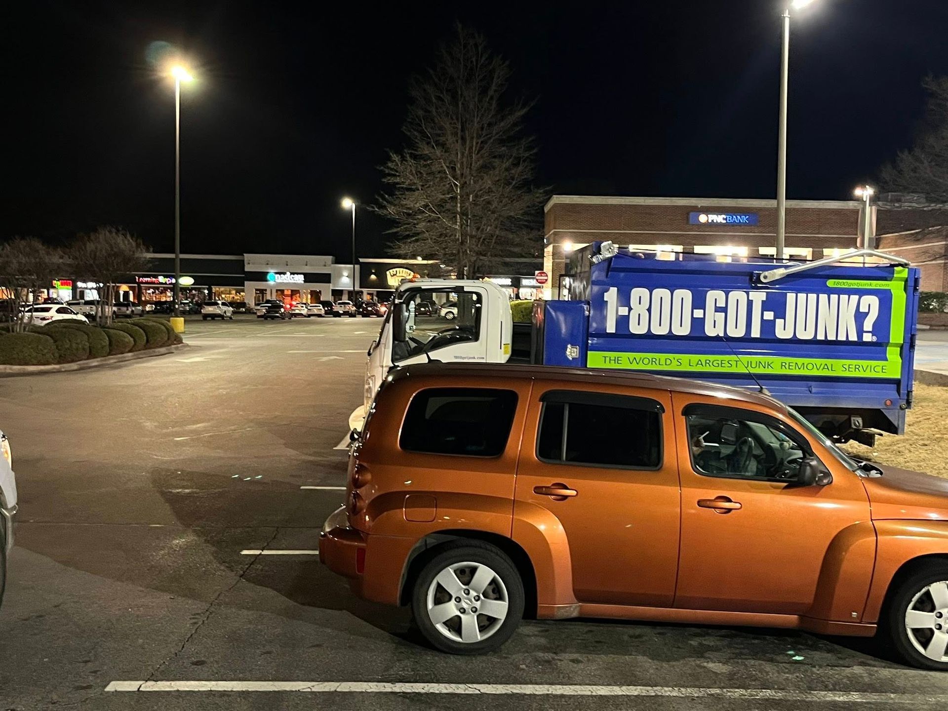 A bright orange SUV parked in a shopping center parking lot at night next to a blue 
