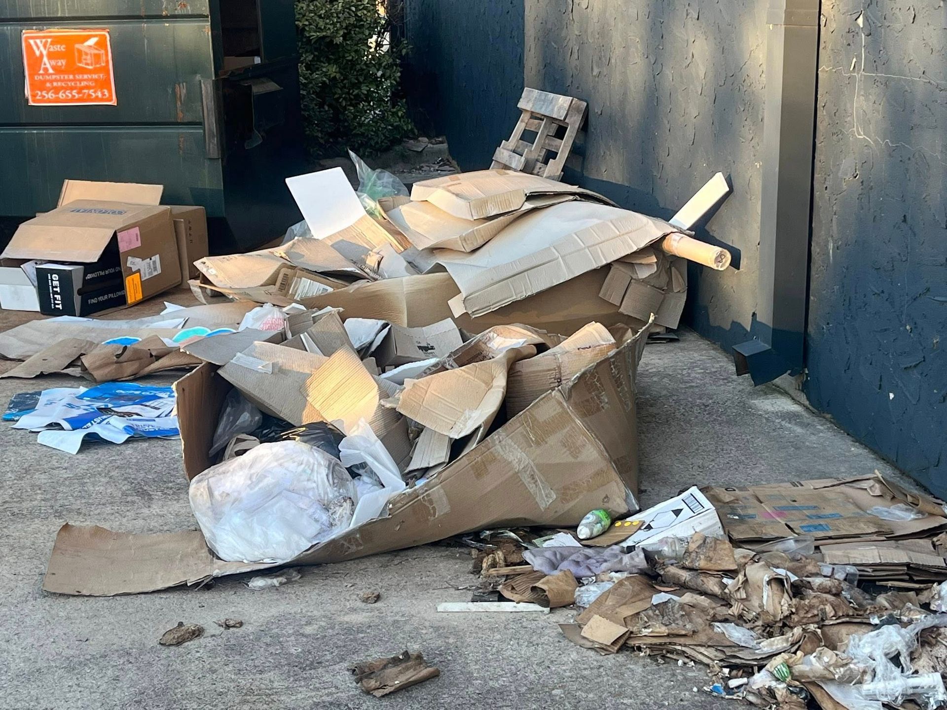 A pile of discarded cardboard boxes and trash on concrete in front of a dumpster and a dark blue wall.