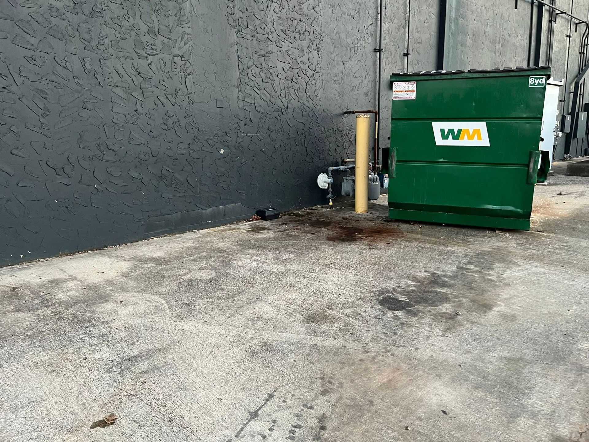 A green Waste Management dumpster stands against a dark gray wall on a paved surface near a gas meter and yellow bollard.