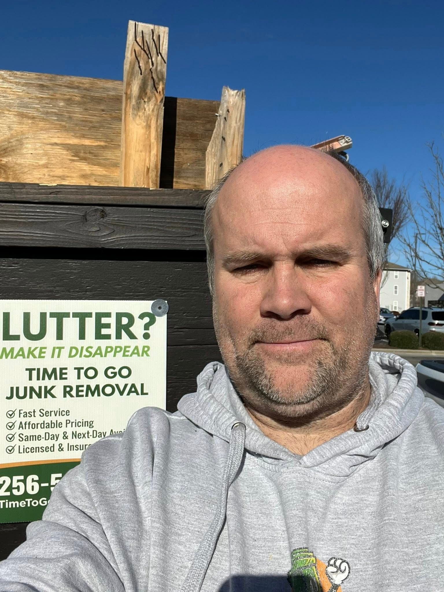 A person stands in front of a dumpster featuring a sign for Time to Go Junk Removal against a bright blue sky.