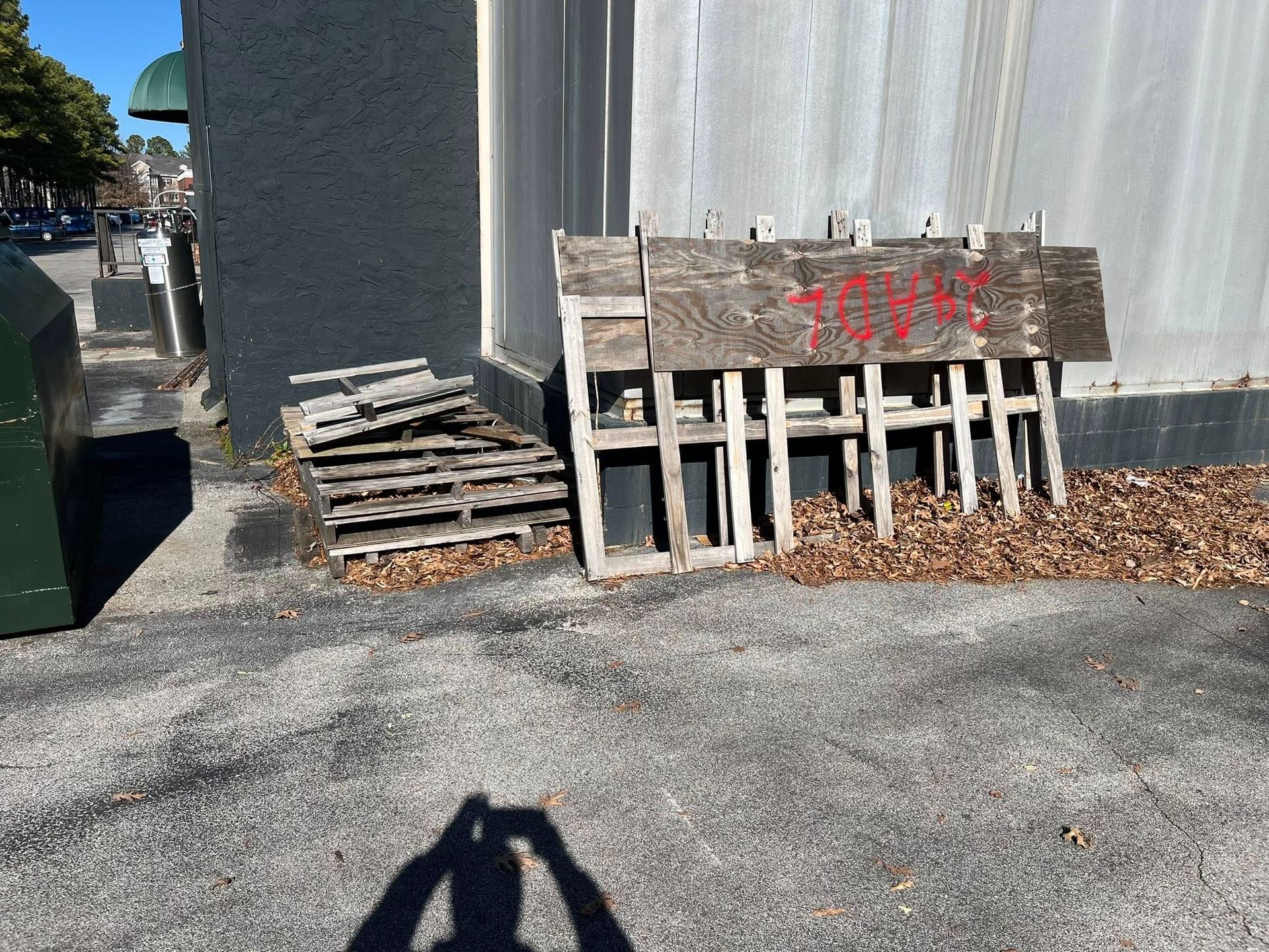 A stack of wooden pallets and a leaning wooden sign with red graffiti markings sit against an outdoor building wall.