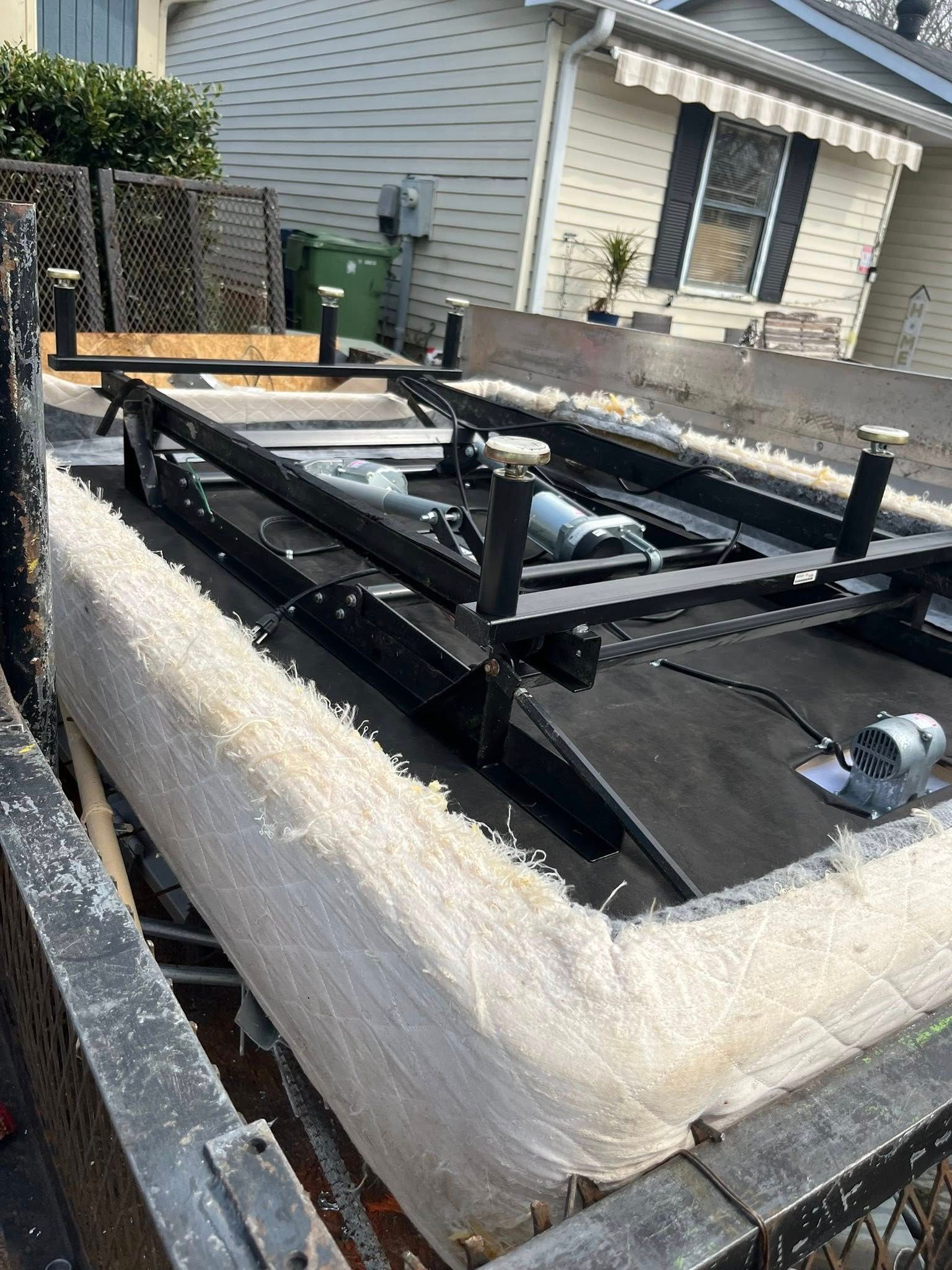 A black metal adjustable bed frame with motors, partially wrapped in plastic, sits on a trailer outside a house.