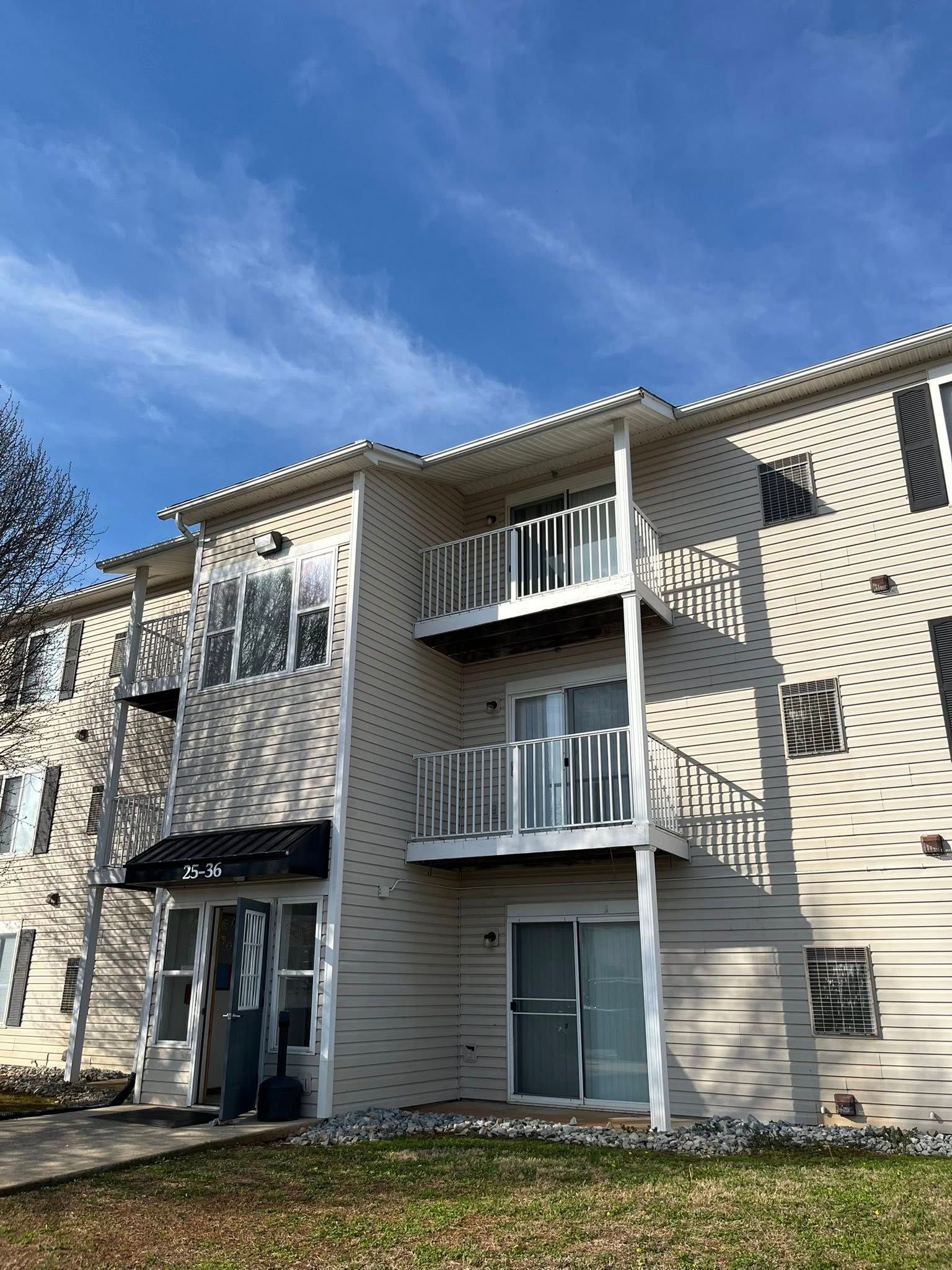 A three-story beige apartment building with white balconies and trim under a bright blue sky.