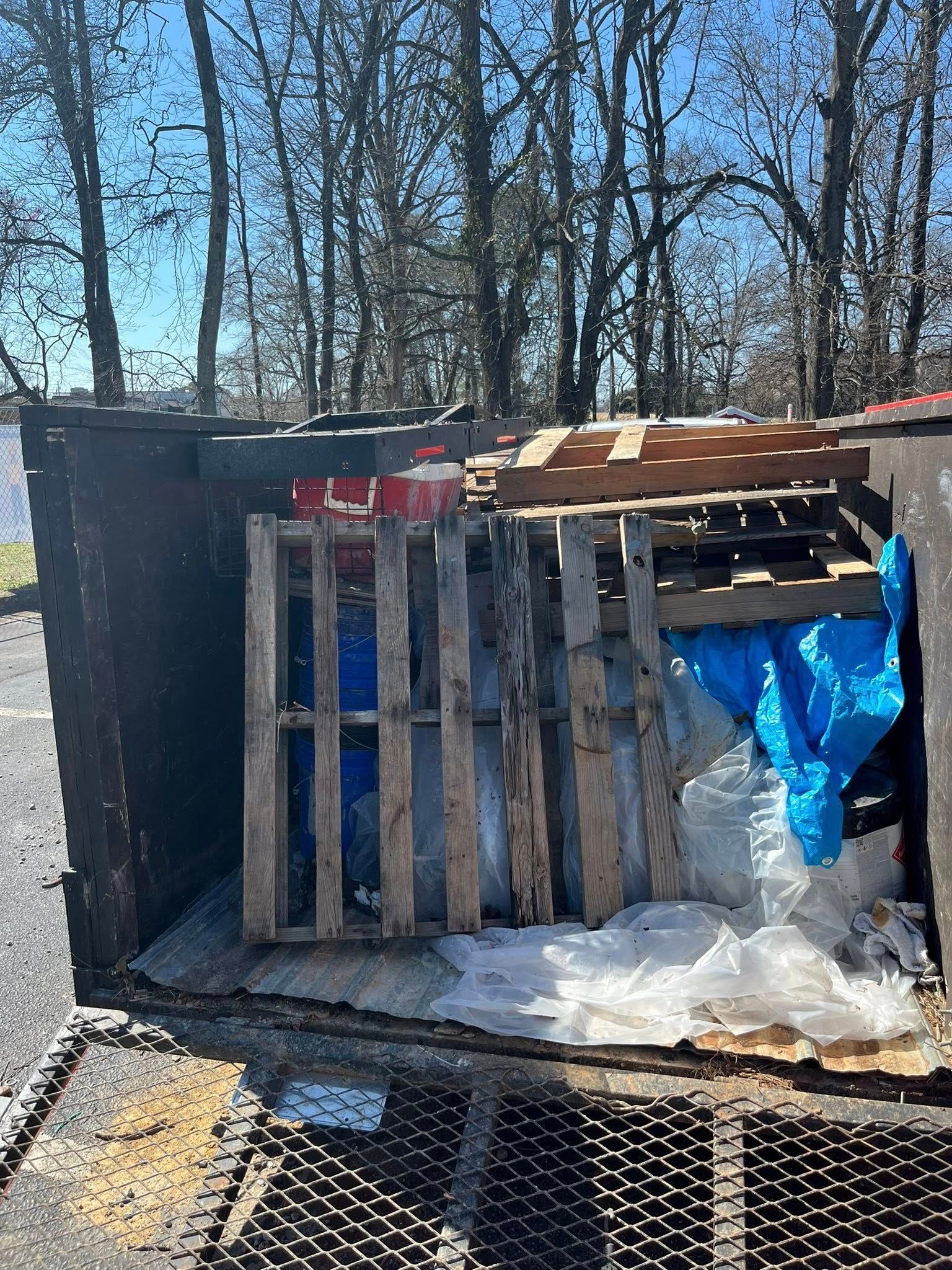 A dumpster containing wooden pallets, a blue tarp, and plastic debris under bright sunlight near a treeline.