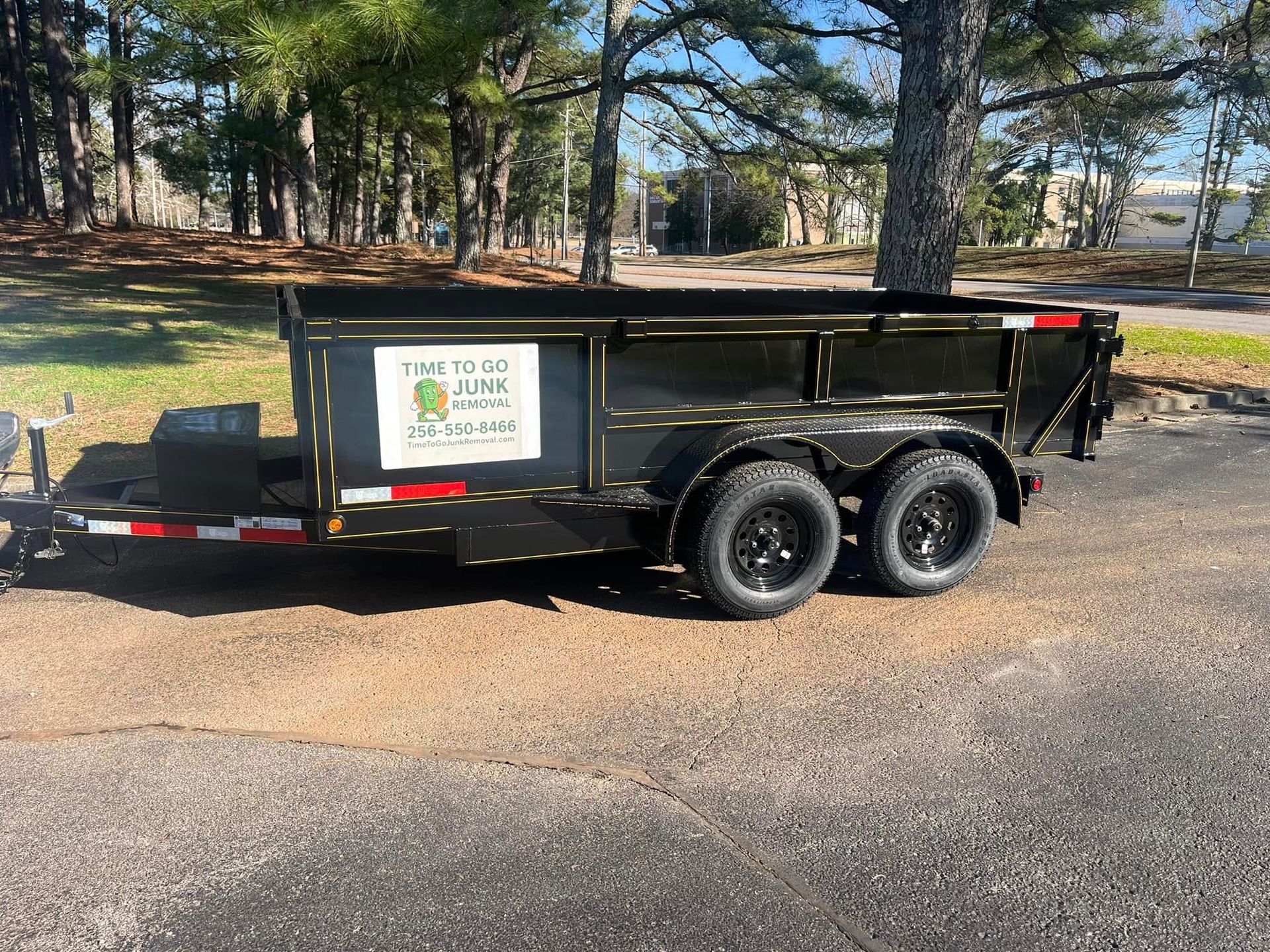A black dump trailer parked on asphalt, featuring a business sign with a logo and phone number on the side panel.