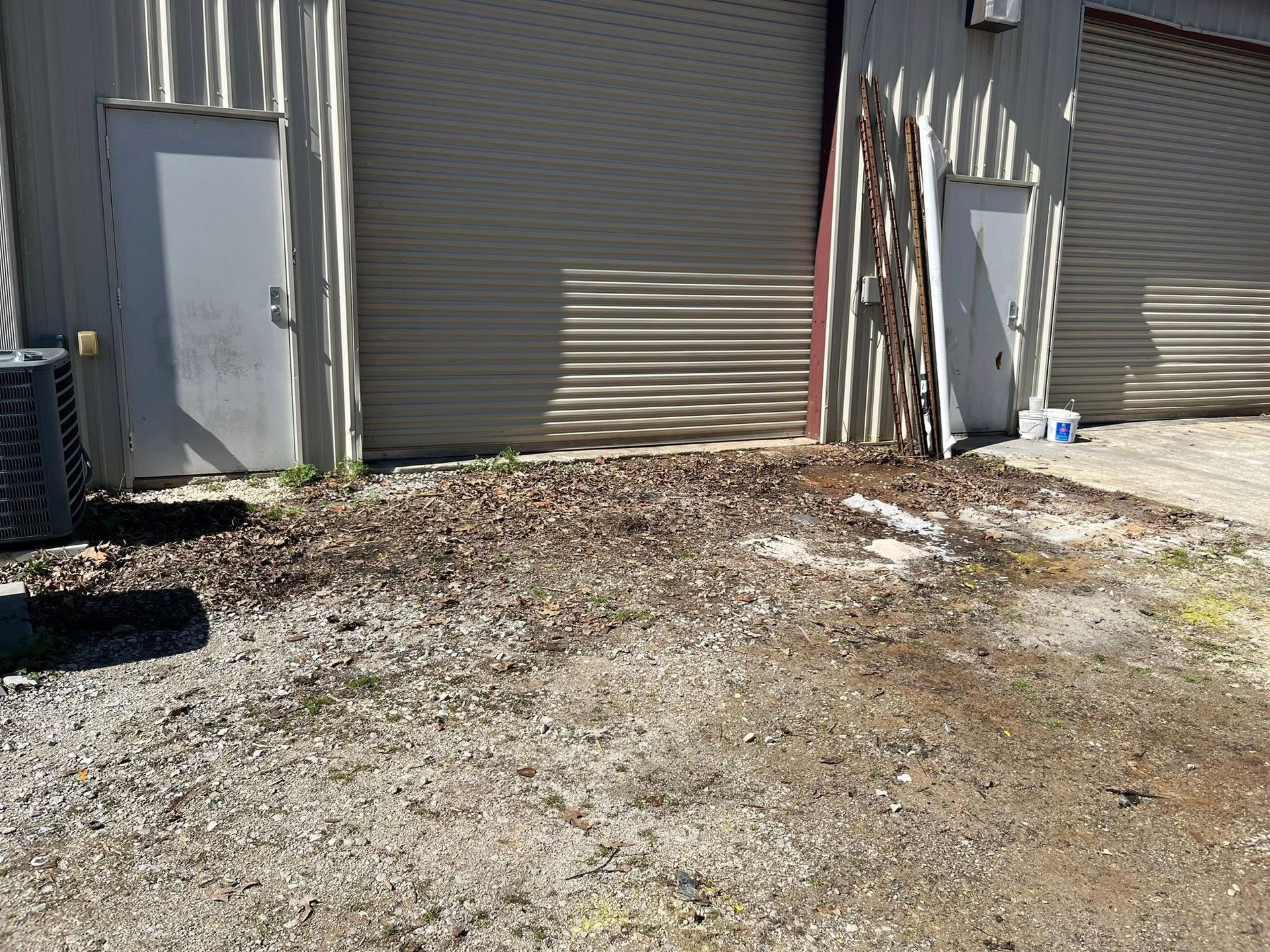 A gravel lot with dry leaves sits in front of two metal warehouse buildings with grey doors and roll-down shutters.