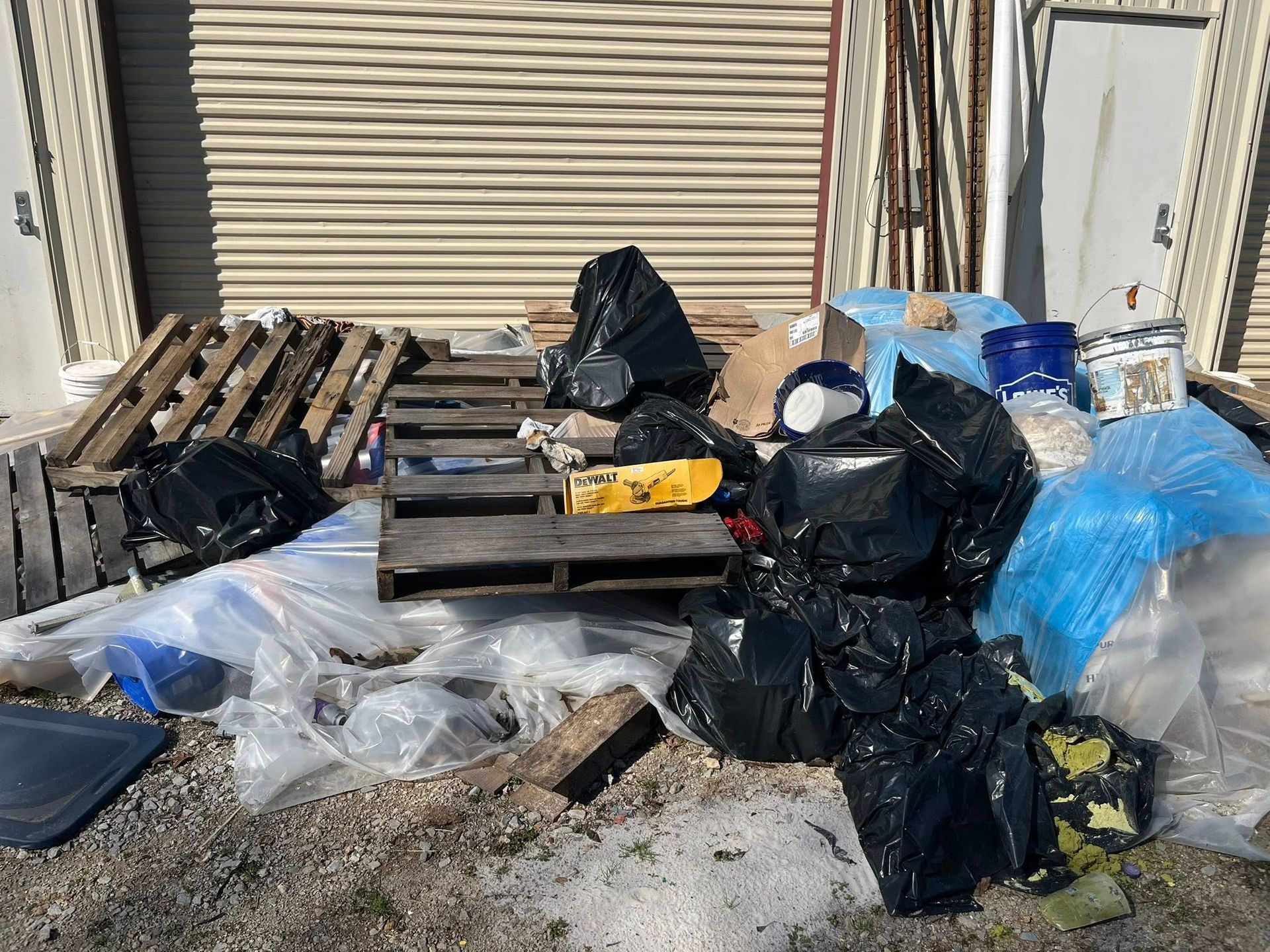 A pile of trash, including black bags, wooden pallets, and plastic sheeting, outside a tan-colored storage building.