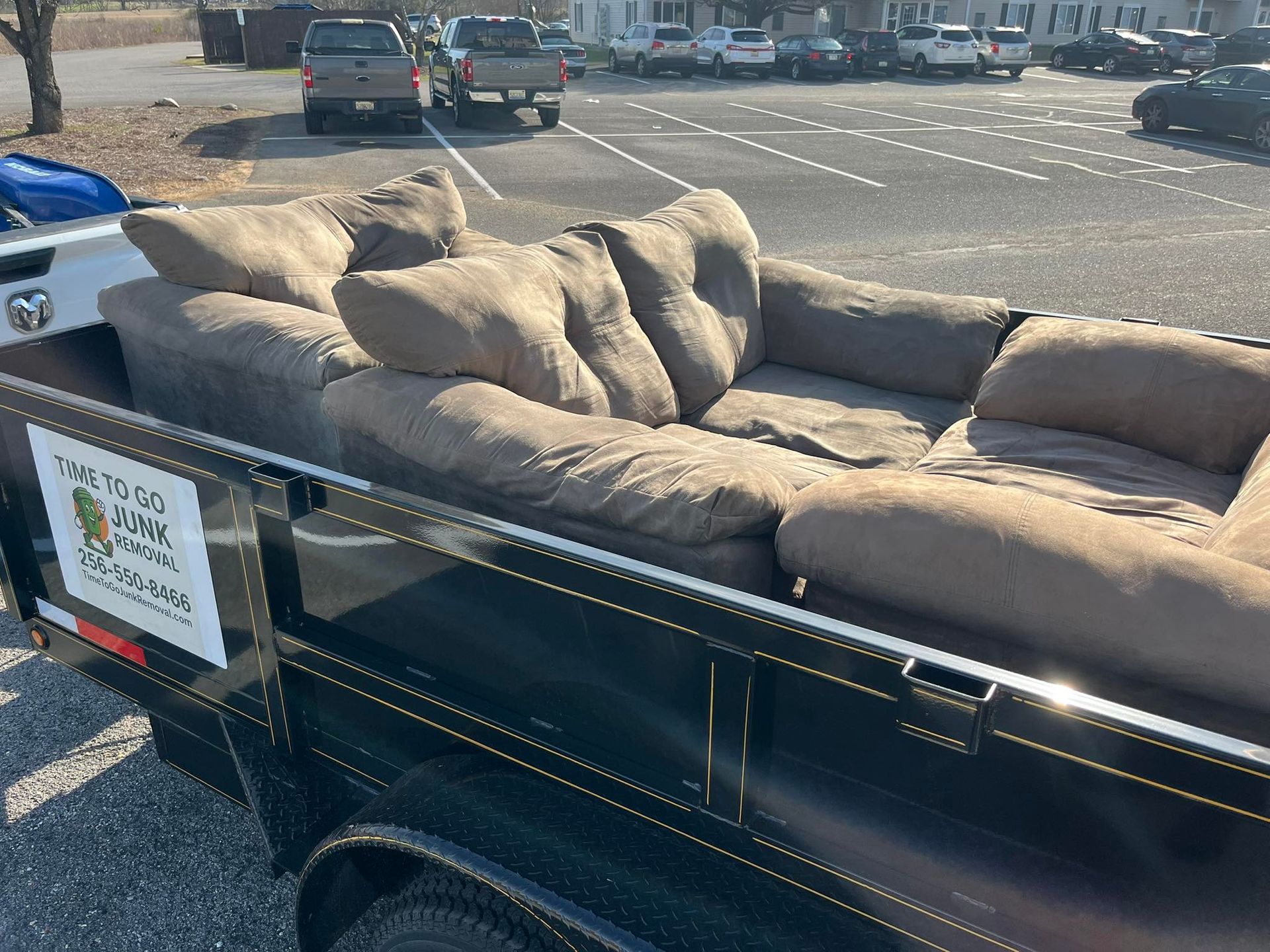 A large, tan sectional sofa sits in a black utility trailer parked in a sunny outdoor lot.