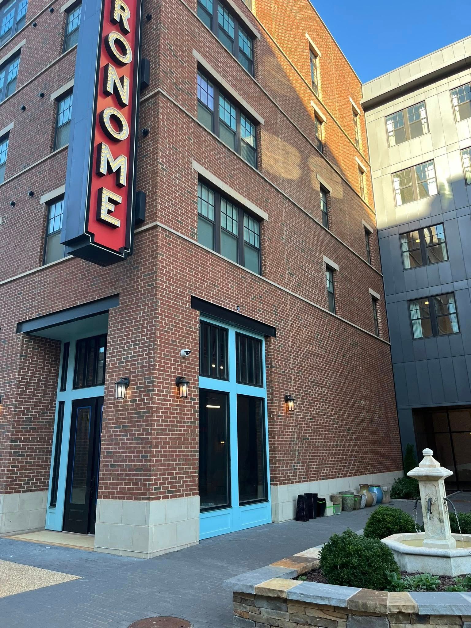 A red brick building corner with a vertical blue “METRONOME” sign, teal window frames, and a small fountain in the plaza.