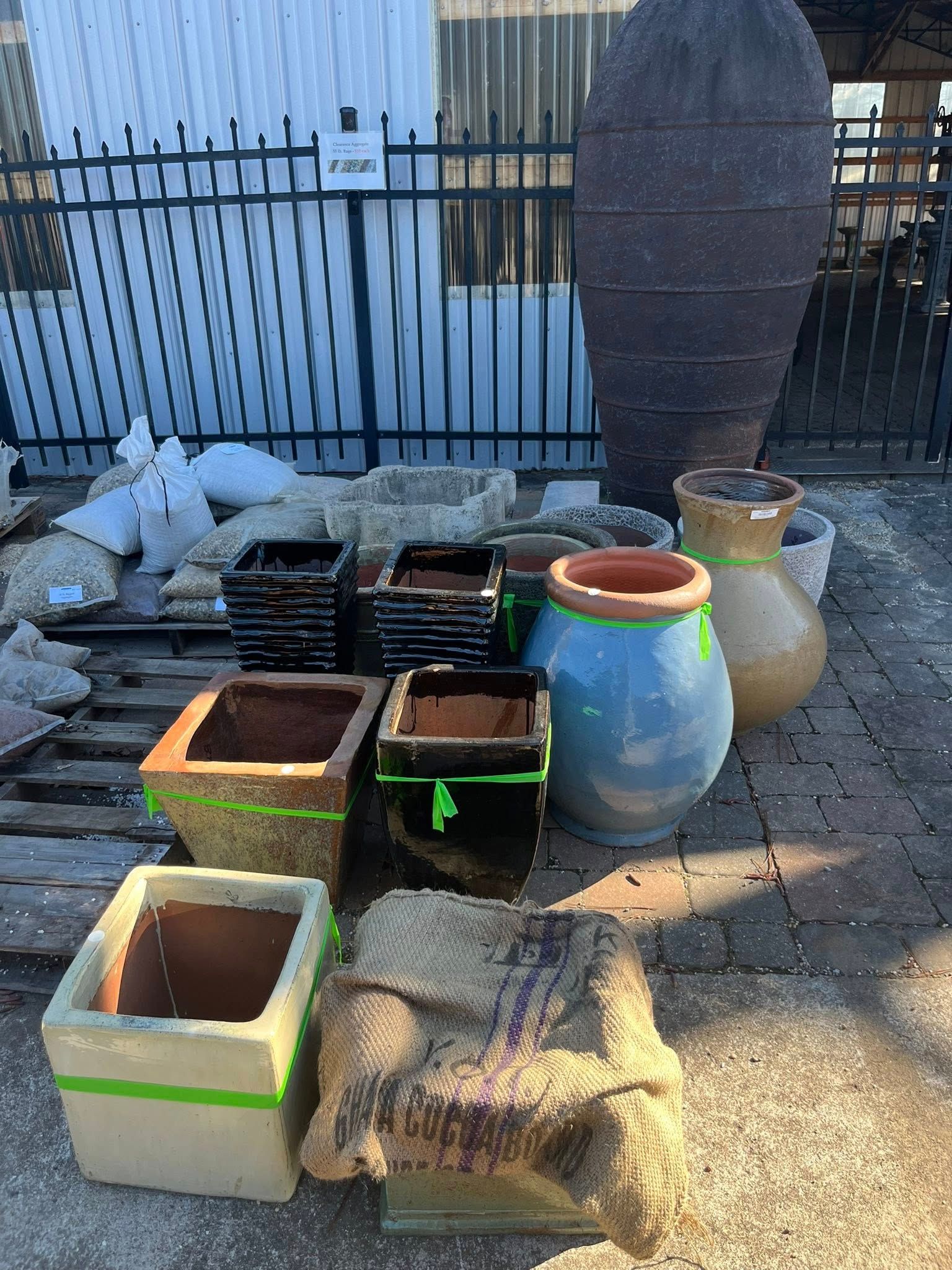 An outdoor collection of stone, ceramic, and burlap-covered planters and pots arranged on a paved patio by a metal fence.