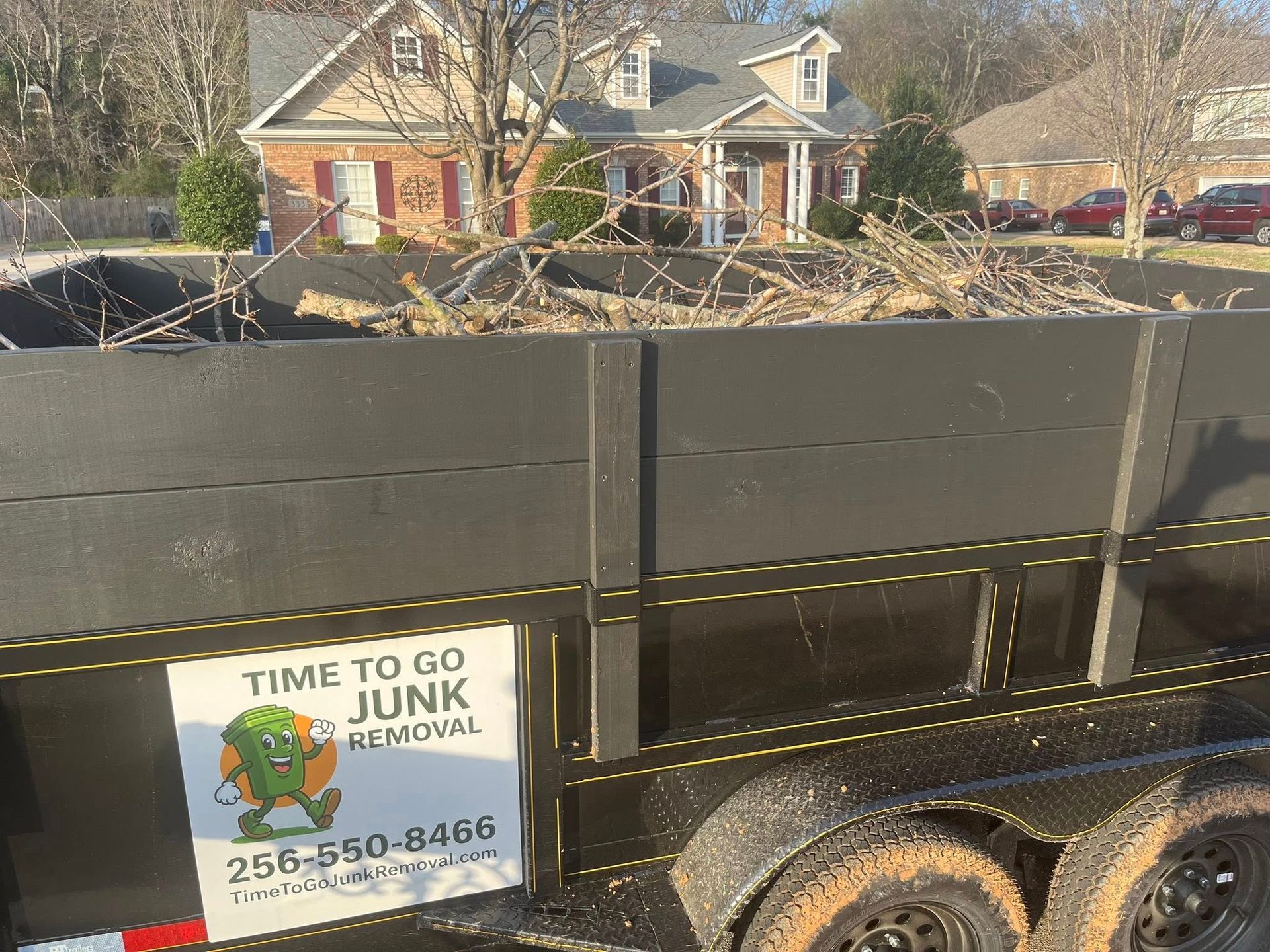 A black junk removal trailer filled with tree branches, featuring the company name and phone number on the side.
