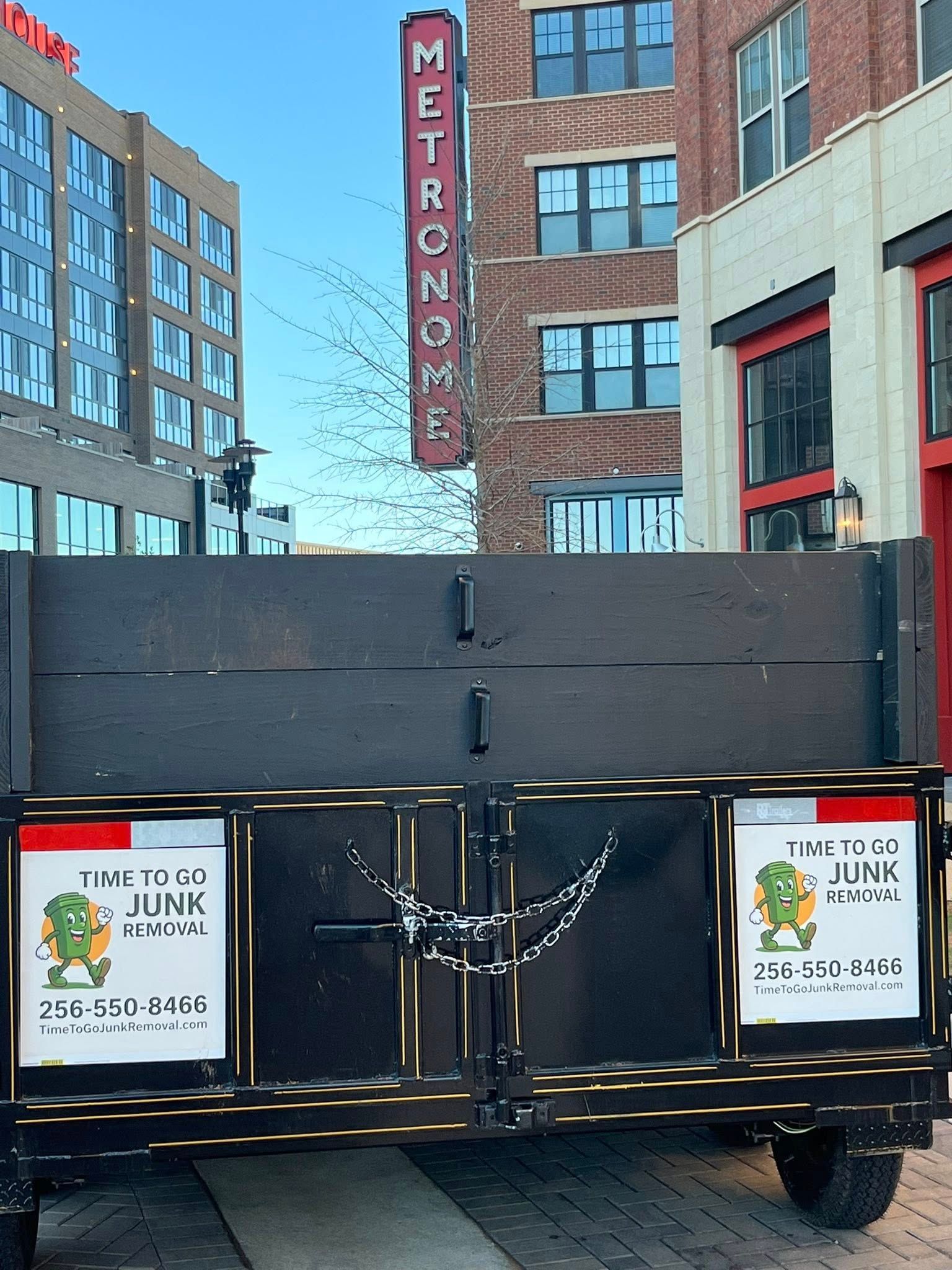 A black junk removal trailer parked on a brick street in front of a brick building with a vertical 