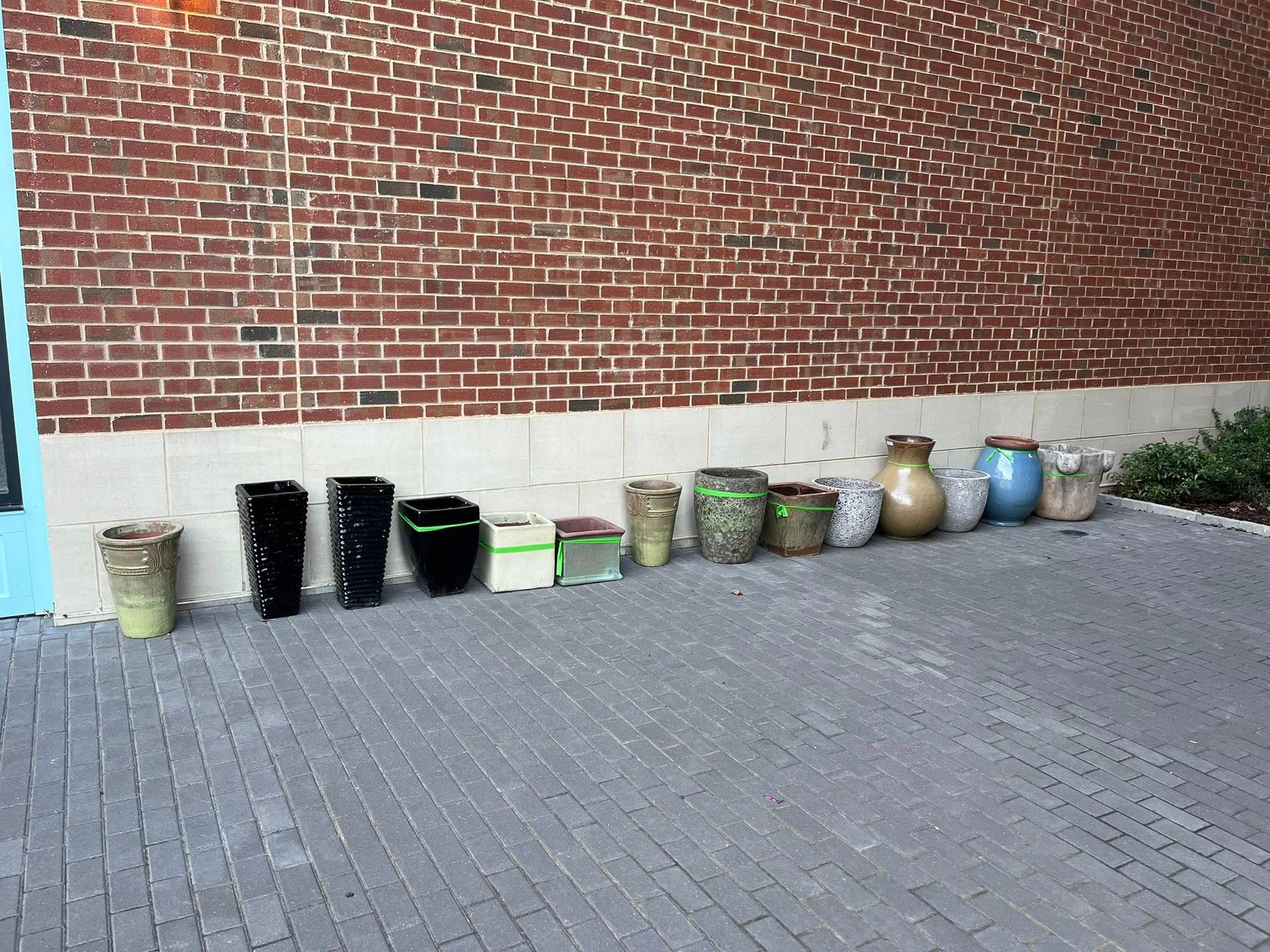 A row of various empty flower pots of different shapes, sizes, and colors lined up against a brick wall on a paved patio.