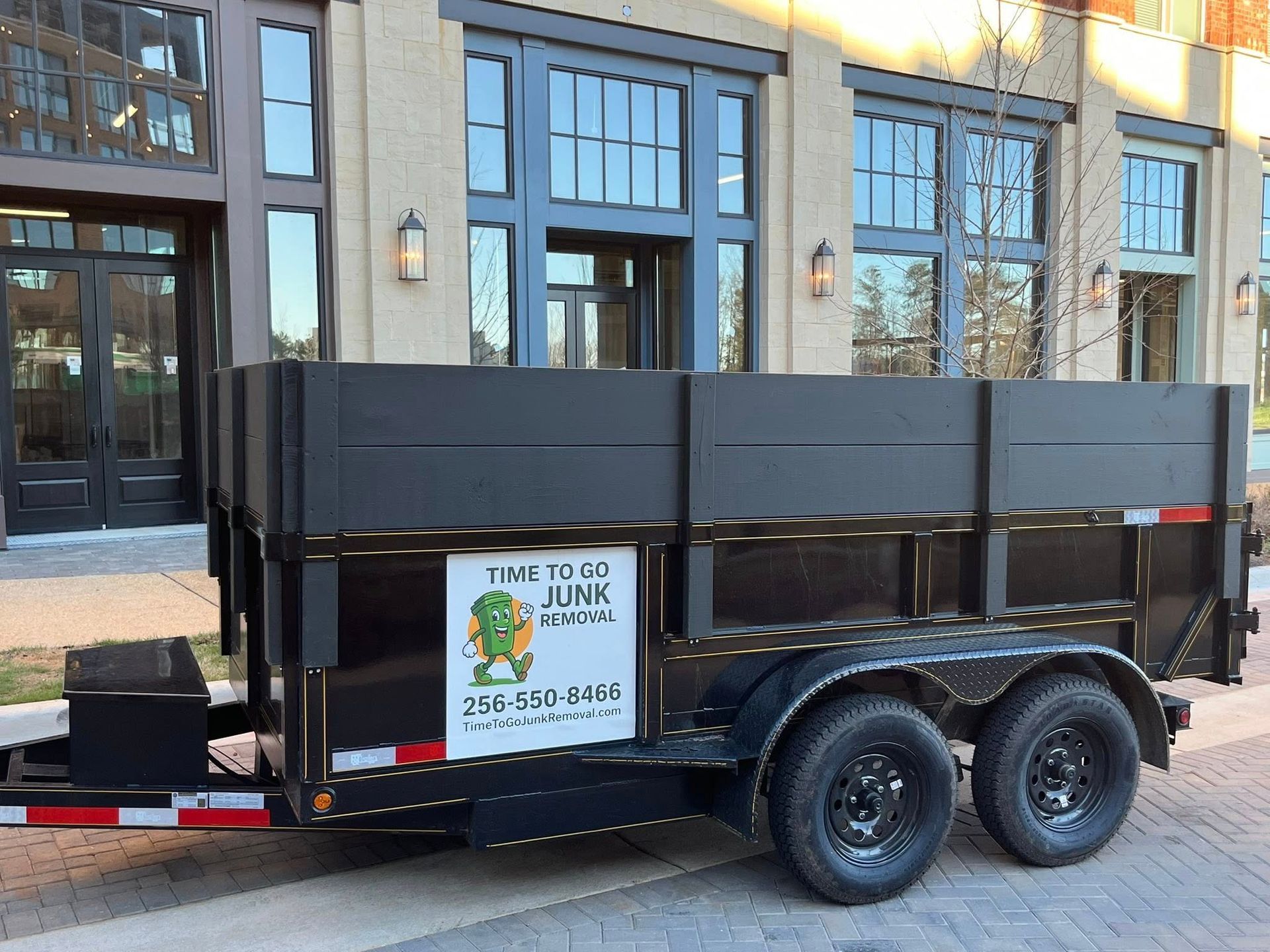 A black junk removal trailer parked on a brick sidewalk in front of a modern multi-story commercial building.