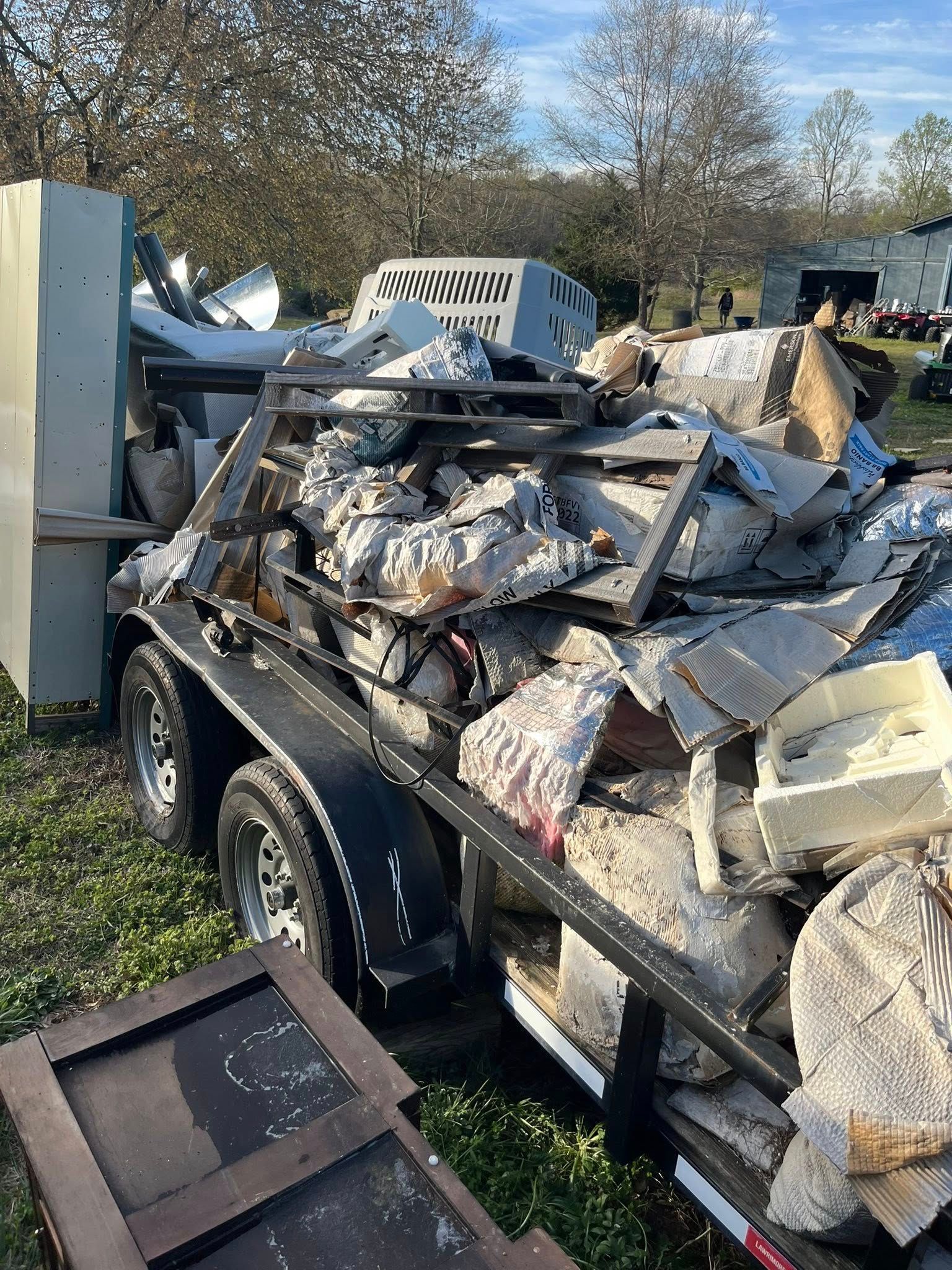 A trailer piled high with construction debris, silver insulation, and metal scrap, parked in an outdoor, grassy area.
