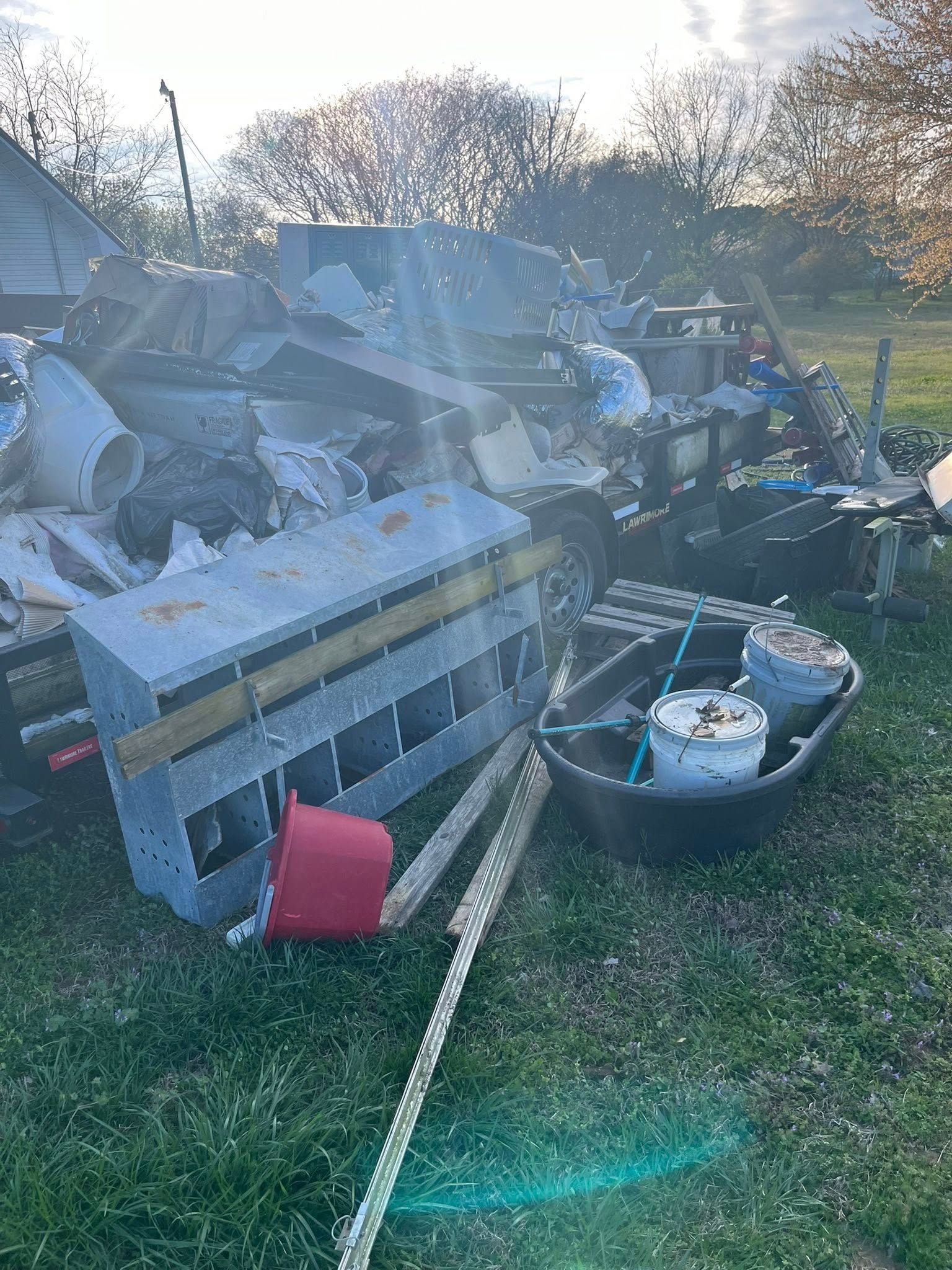 A messy pile of junk, including a gray metal nesting box, a red bucket, and a black plastic tub, sitting on grass.