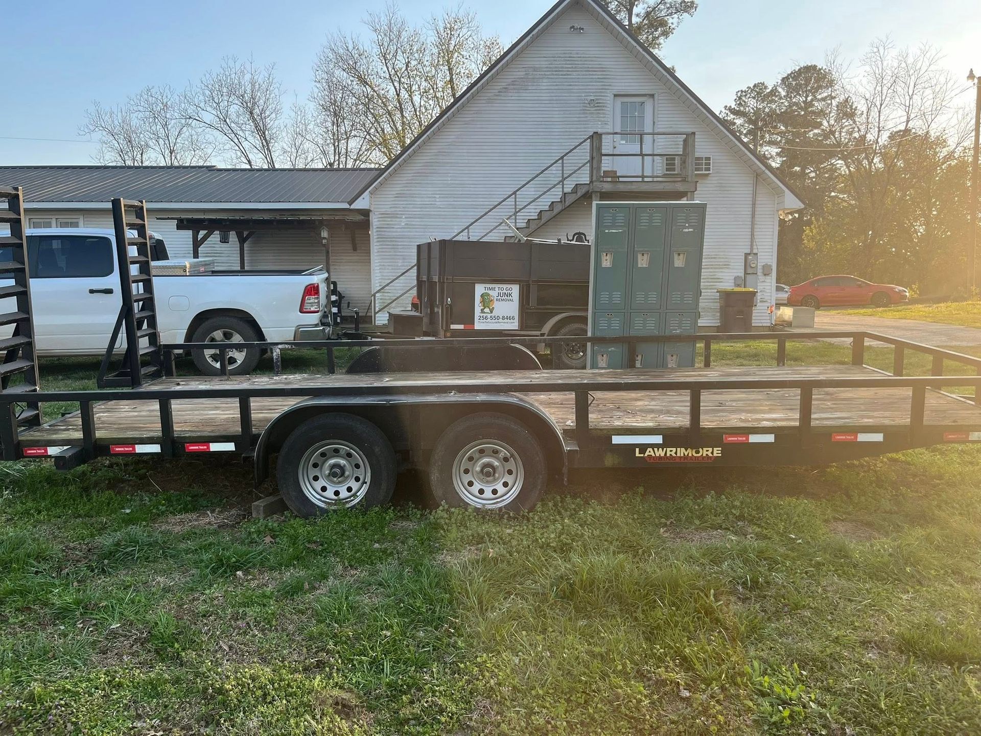 A flatbed utility trailer parked in a grassy field in front of a white building with an exterior staircase.