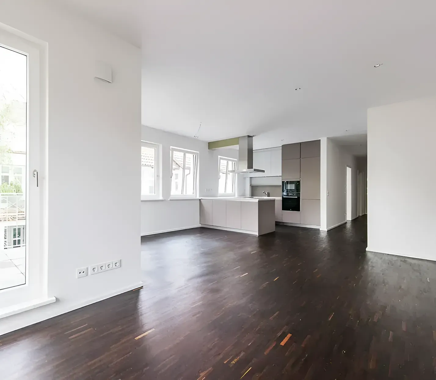 An open-concept living area with dark wooden floors, white walls, and a minimalist kitchen in the background.