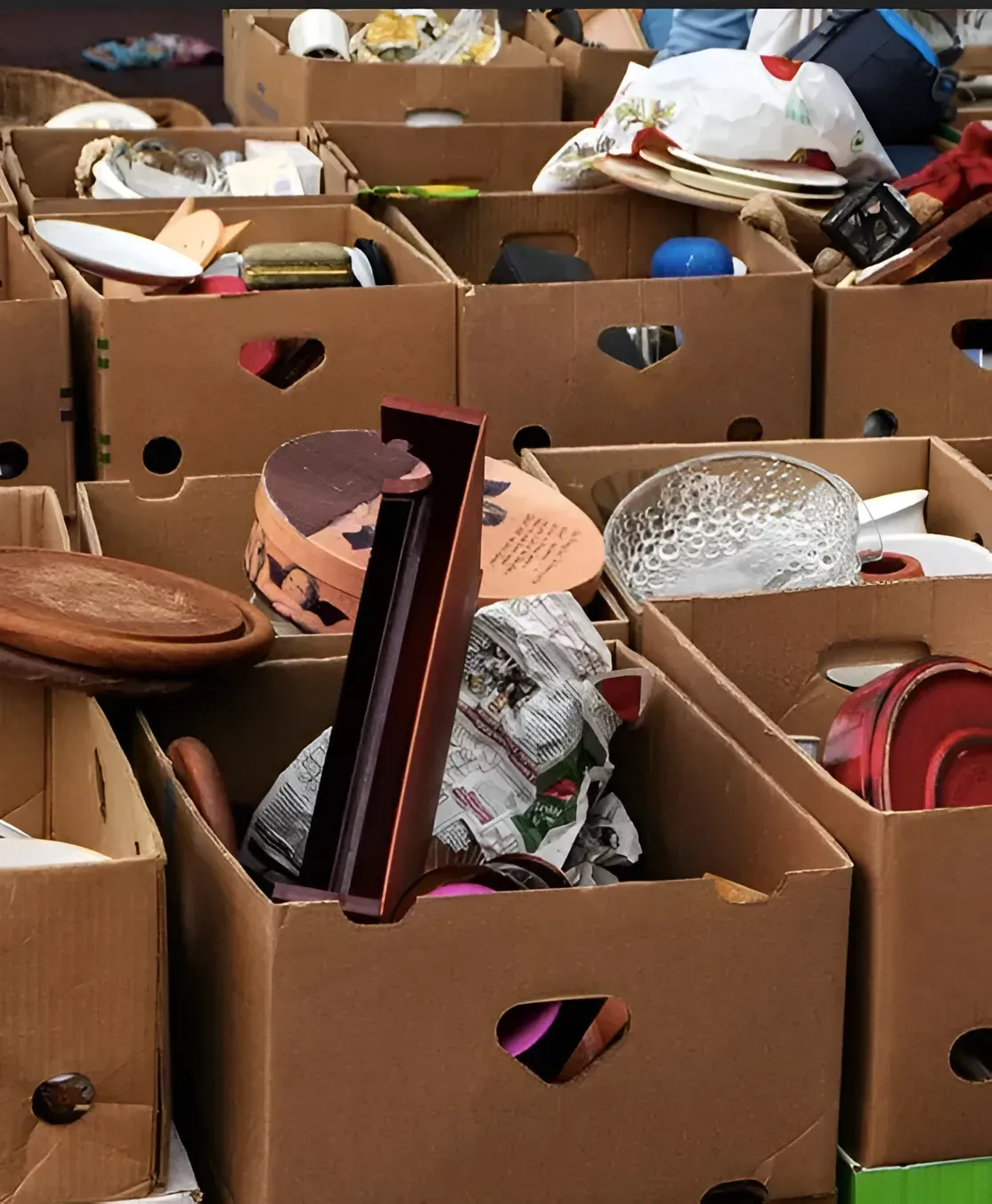 Cardboard boxes filled with assorted household items like bowls, plates, and miscellaneous clutter at a yard sale.