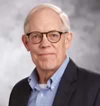 Elderly man with glasses, wearing a blue collared shirt under a dark blazer, smiles at the camera.