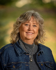 Woman with wavy gray hair smiles, wearing a denim jacket and gray turtleneck outdoors.