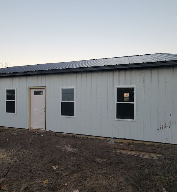 A white house with a black roof and a white door is sitting in the middle of a dirt field.