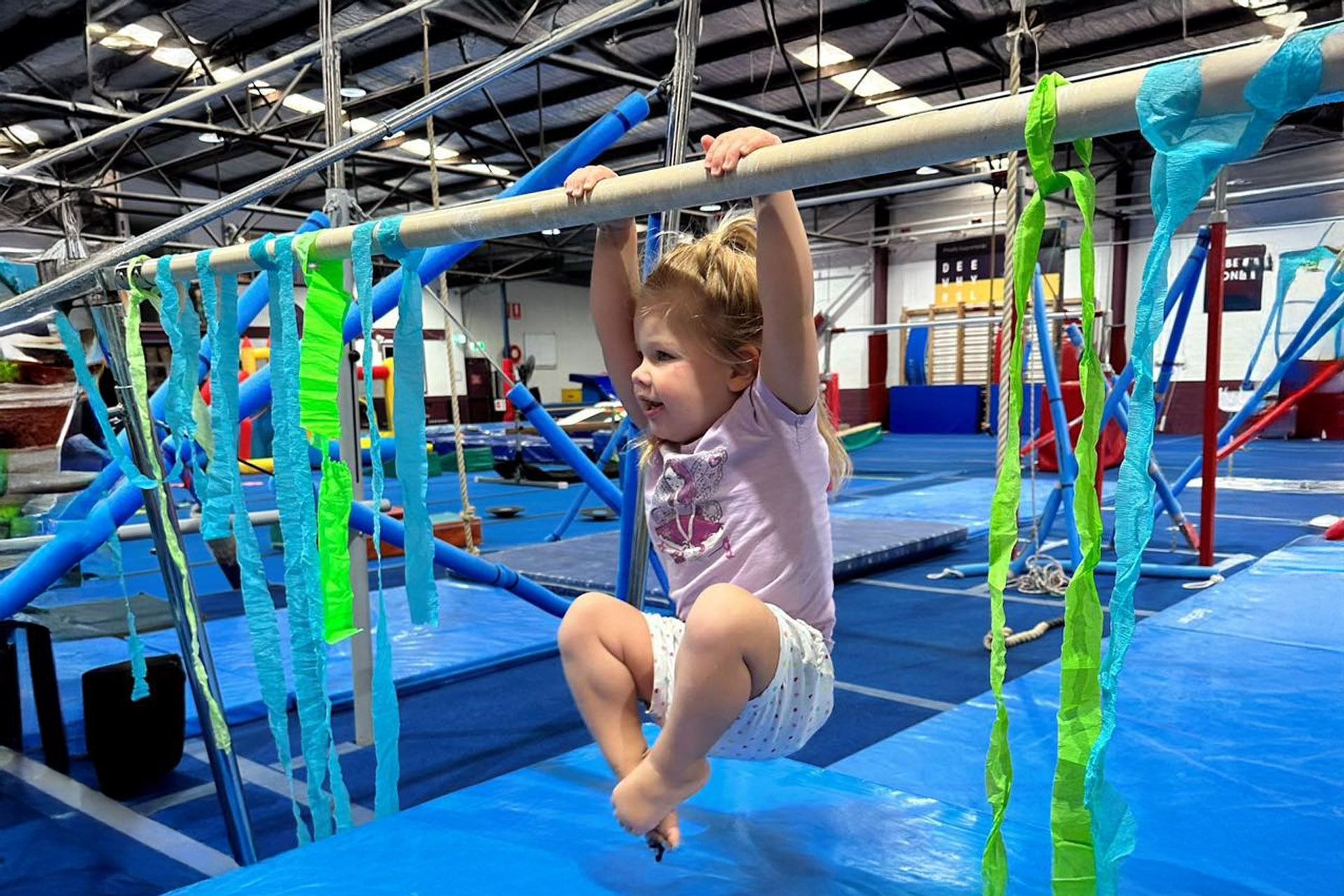 A little girl is hanging on a bar in a gym.