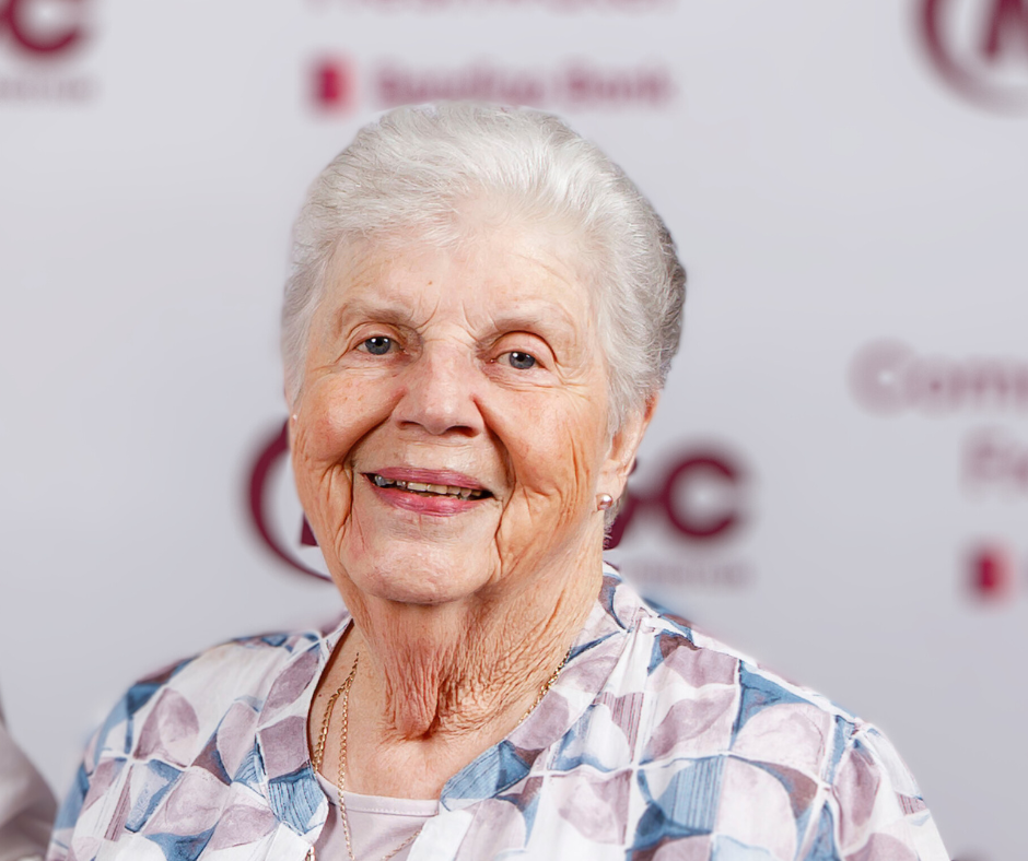 An elderly woman is smiling for the camera in front of a blurred background.