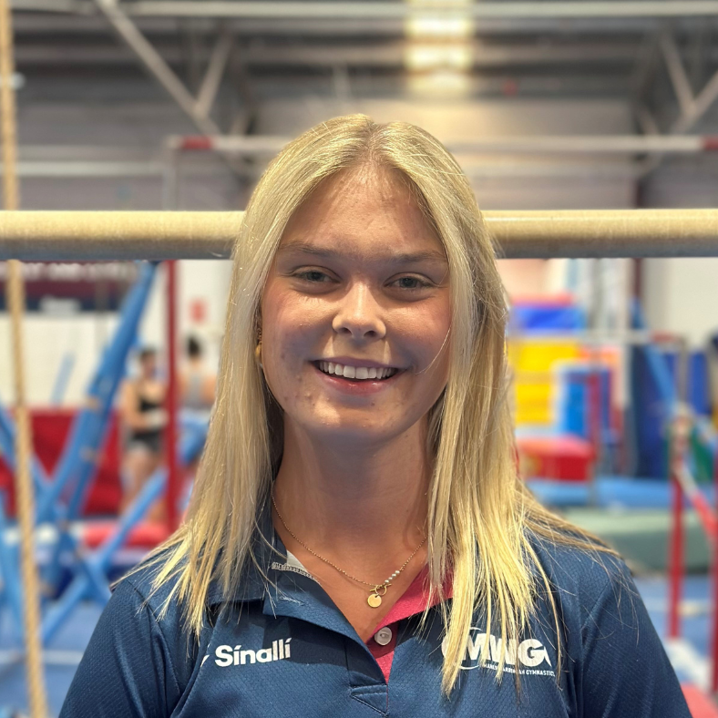 A woman is smiling in front of a balance beam in a gym.