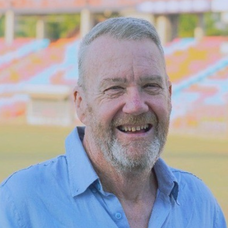 A man with a beard and a blue shirt is smiling in front of a stadium.