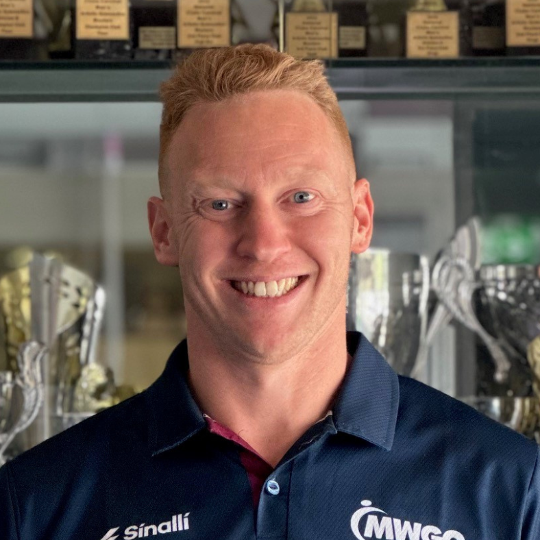 A man wearing a sinalli shirt is smiling in front of trophies