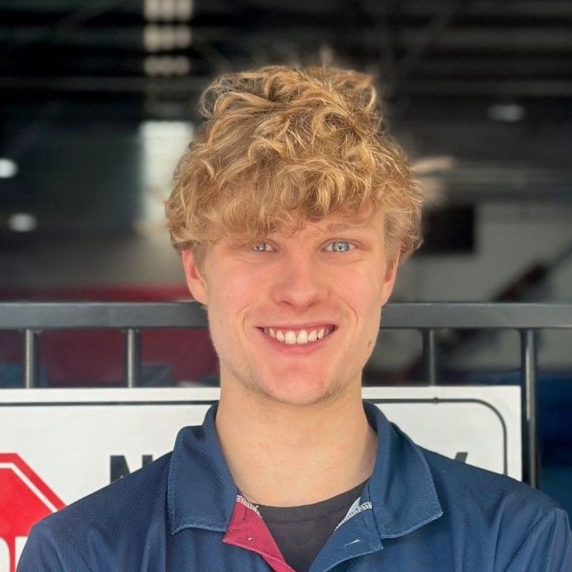 A young man with curly hair is smiling in front of a sign.