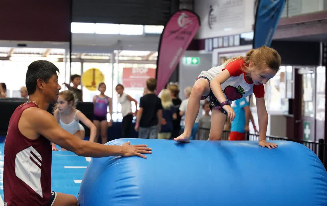 A man is helping a little girl climb on a blue inflatable cylinder.