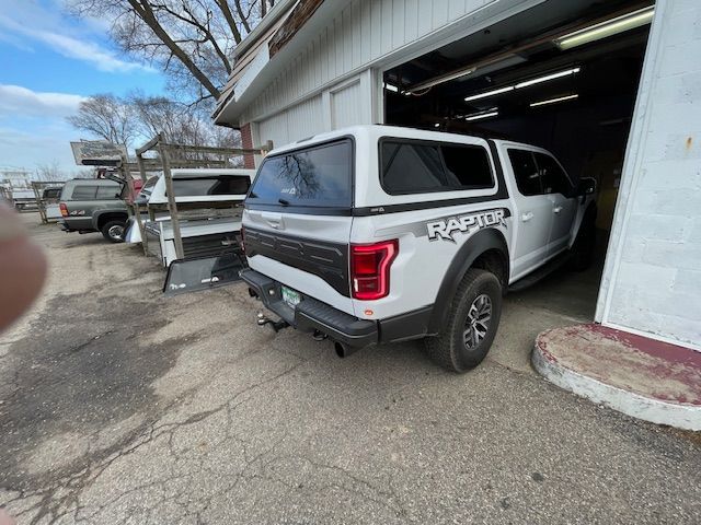 White Ford Raptor truck with a camper shell parked outside a garage.