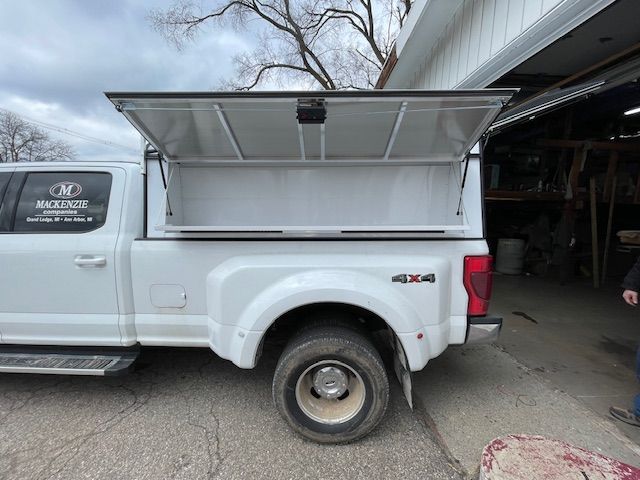 White Ford truck with a white utility bed topper open, parked outside a building.