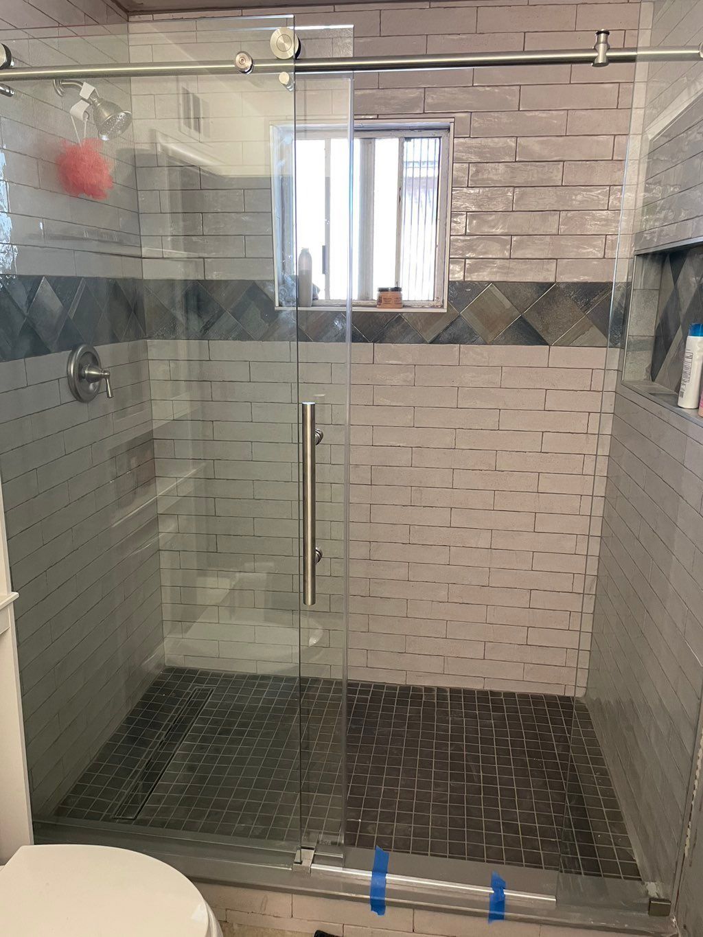 Glass shower with tiled walls and floor, gray, blue, and white tones, a window, and a sliding door.