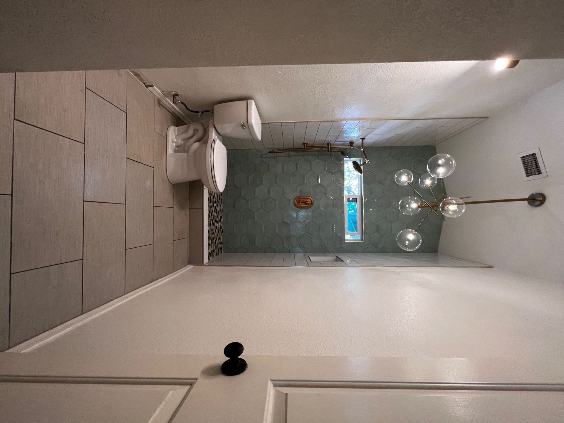 Bathroom with a toilet, shower, and globe chandelier. Gray and white tiles.