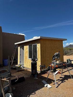 Construction of a wooden shed outdoors with a saw and ladder present under a blue sky.
