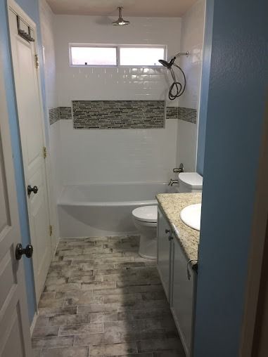 Small bathroom with white and stone tile, white tub, toilet, vanity, and door.
