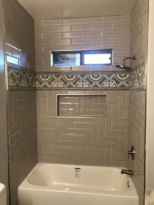 Gray tiled shower with bathtub, decorative border, and small window.