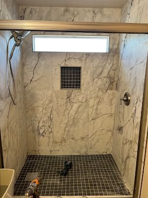 A tiled shower with marble-look walls and a dark mosaic floor, featuring a recessed shelf and overhead light.