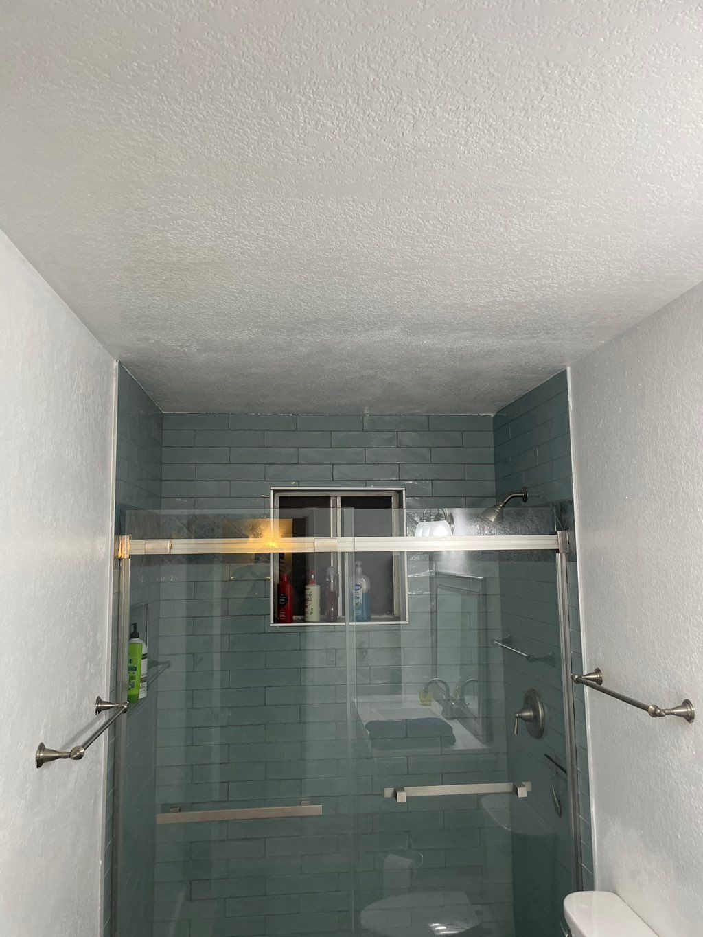 Bathroom with blue tiled shower, glass doors, and a small window. Towel racks and toilet visible.