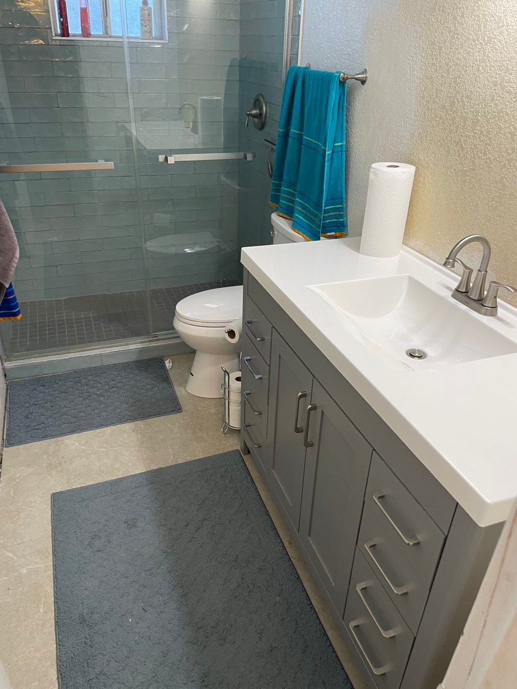 Bathroom with gray vanity, white sink, toilet, glass shower, blue towels, and rugs.