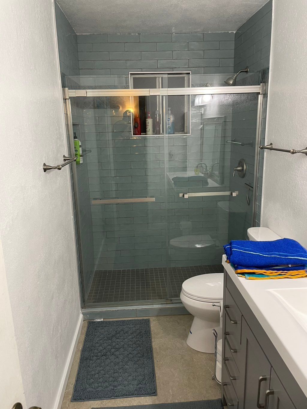 Small bathroom with blue tile shower, toilet, vanity, and towels.