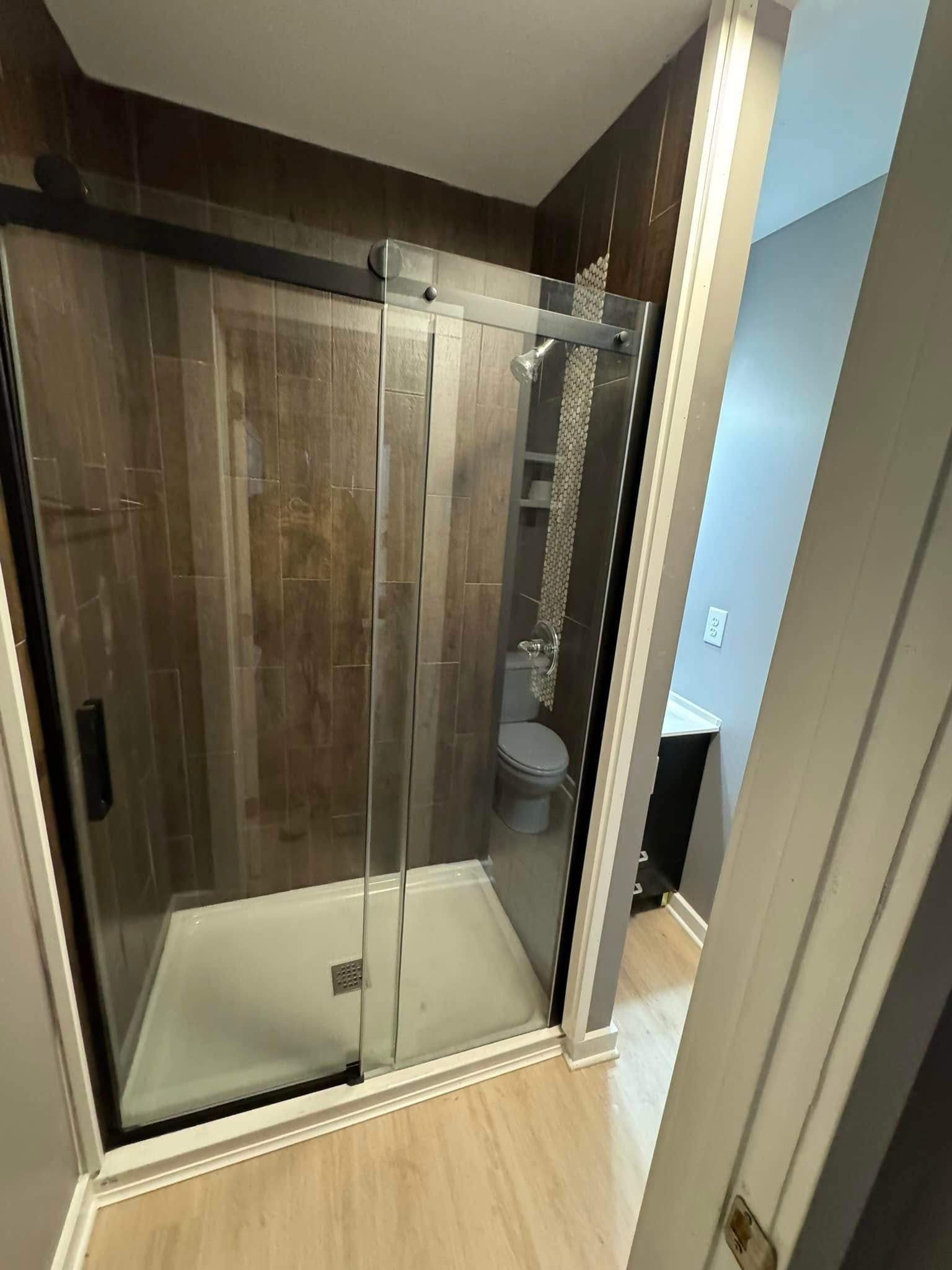 Bathroom with a modern glass shower, brown tiled walls, and a toilet visible in the background.