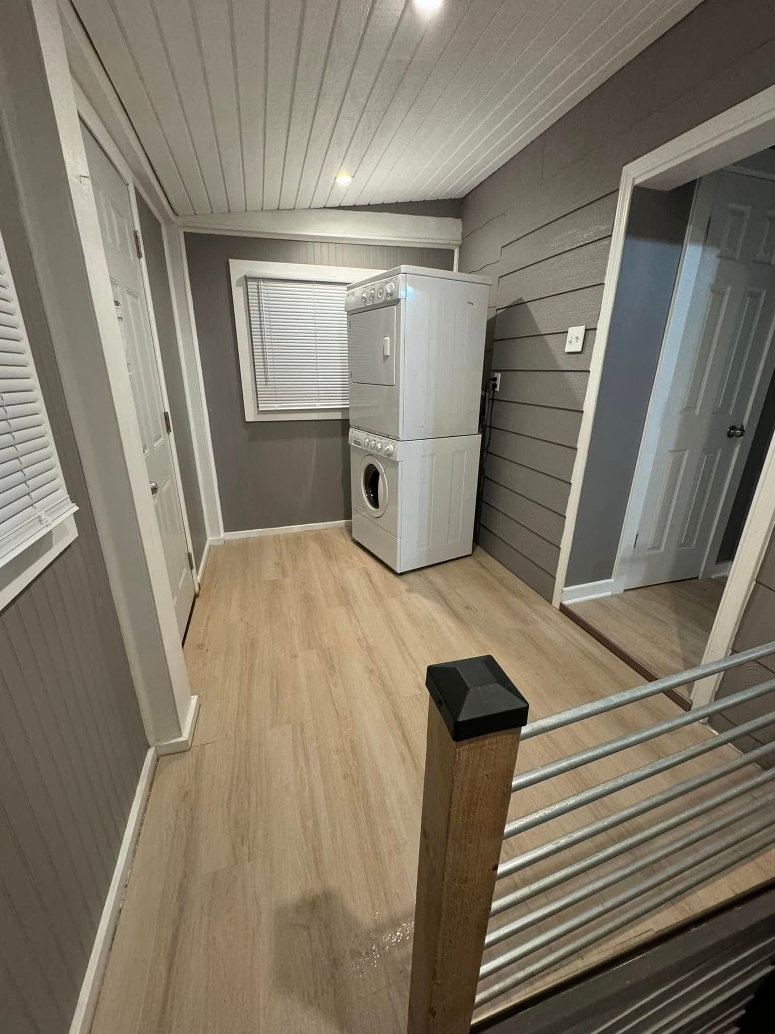Laundry room with stacked washer and dryer, wood-look flooring, and gray walls. Sunlight streams through the window.