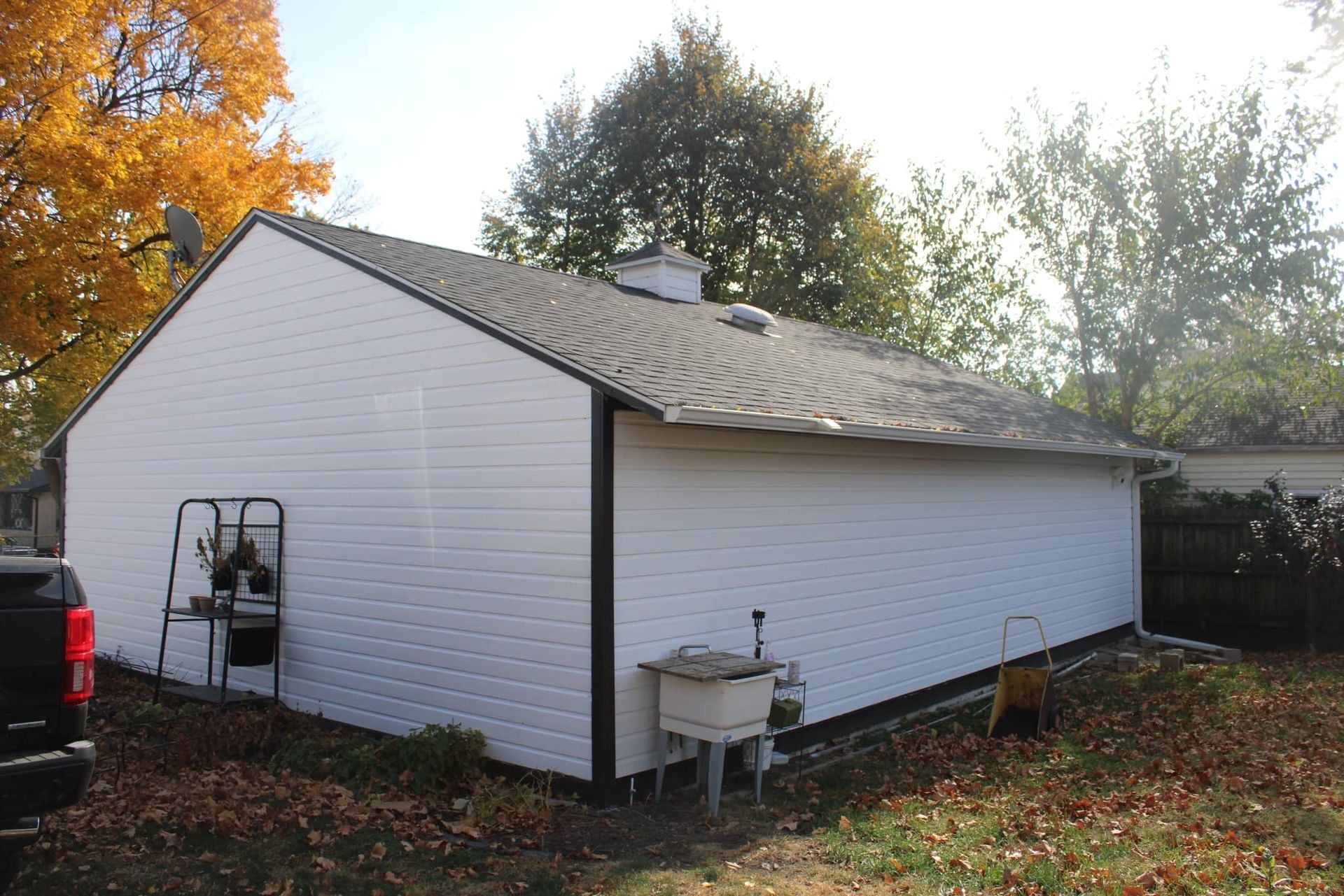 A white shed with a dark roof and trim stands in a yard with fall foliage and an outdoor sink.