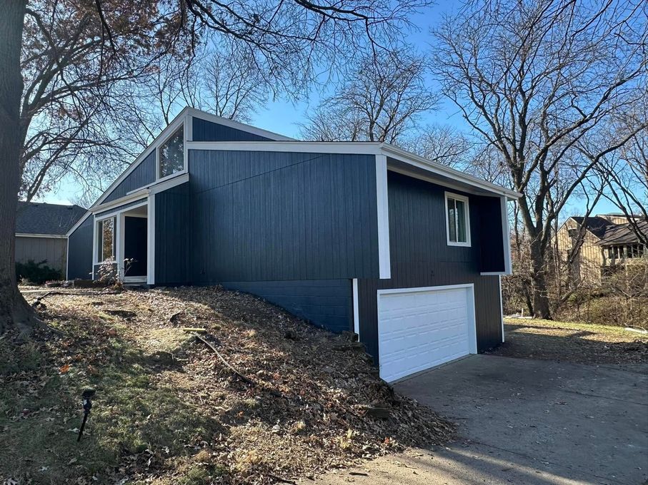 A two-story house with blue siding and white trim sits on a slight hill. The driveway leads to a white garage door.