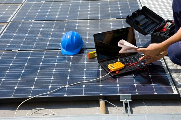 Persona inspeccionando paneles solares con una computadora portátil y un multímetro en un tejado, usando un casco azul.